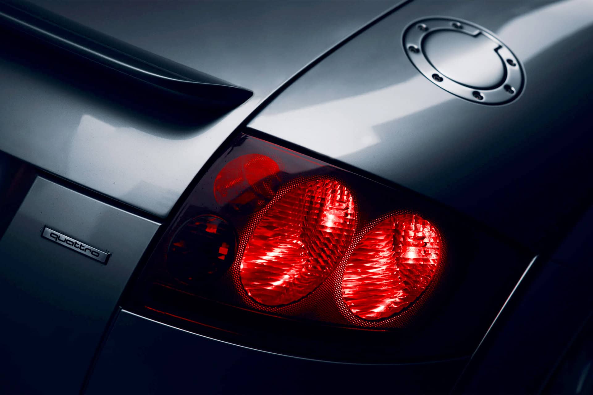 A sleek silver luxury car rear close-up showing illuminated red tail lights, rear design details, and the "quattro" badge, emphasizing high-performance automotive style and modern engineering.