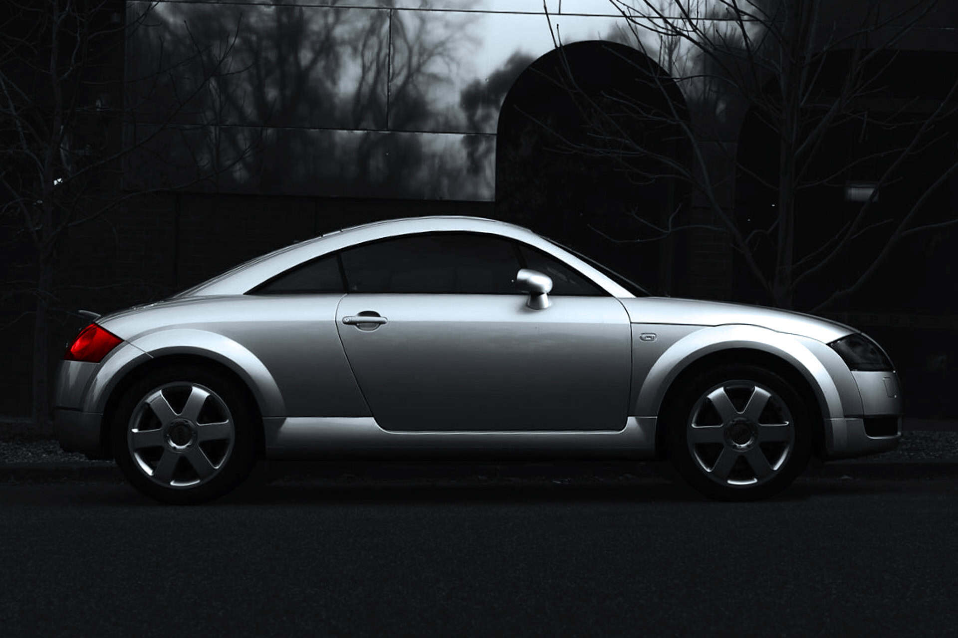 Silver Audi TT sports car parked on dark street at dusk with trees and buildings in background.