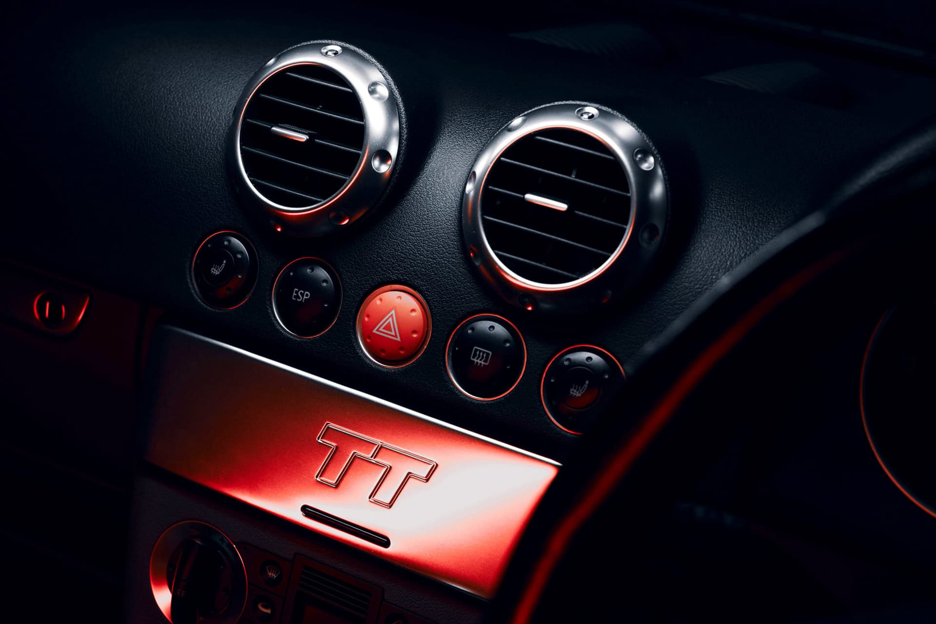 Vivid picture of a luxury car's dashboard featuring circular air vents, control buttons, and ambient red lighting with a TT logo, emphasizing sleek design and modern automotive interior.
