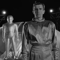 Blonde man in metallic sci-fi costume standing in front of spaceship, black and white, futuristic performance, space-themed fashion, science fiction costume, concert stage, I AM GORT.
